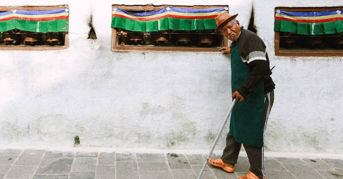 Un homme âgé en tenue traditionnelle marche avec une canne à côté de roues de prière colorées dans une rue.