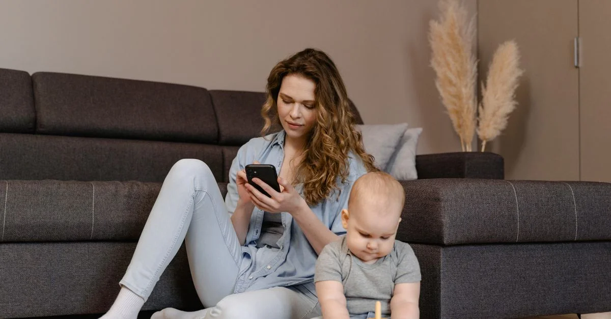 Une jeune mère s'assoit avec son enfant jouant sur le sol, concentrée sur son téléphone.